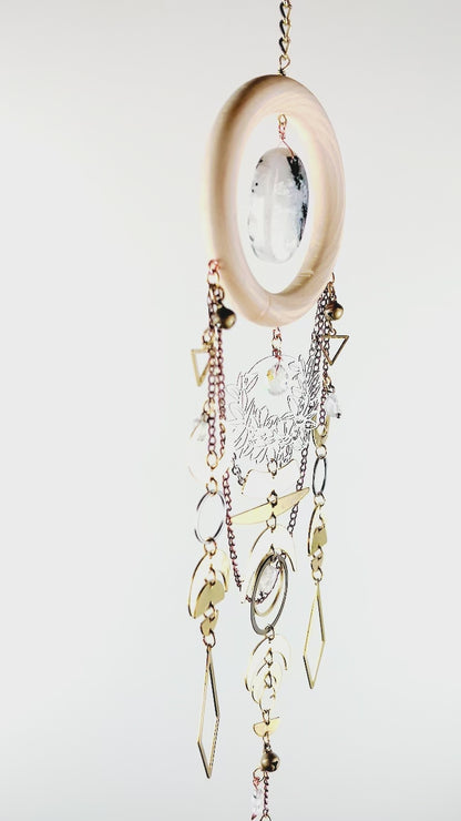 A piece of crystal wall art slowly spins in front of a white surface. This piece includes vertically hanging patterns of gold colored and silver colored metal charms such as: triangles, bells, chain, a flower wreath, circles, crescent moons, half moons, diamond shapes, and a snake Charm.