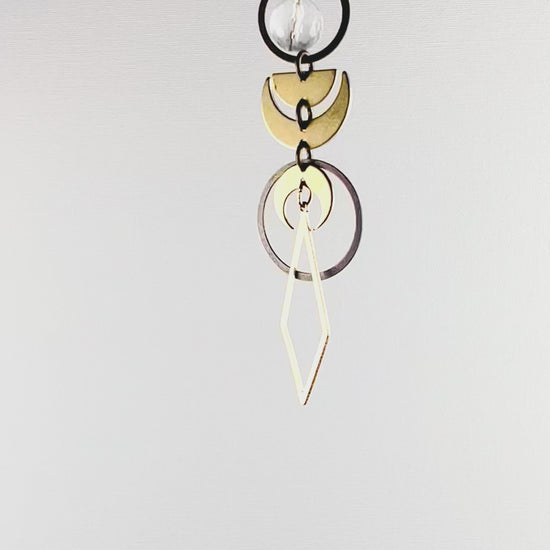 A slowly rotating piece of crystal rearview mirror art made up of connected gold colored metal components such as Rings, crescent moons, half moons and diamond shapes with a faceted clear quartz crystal bead hang in front of a white wall.