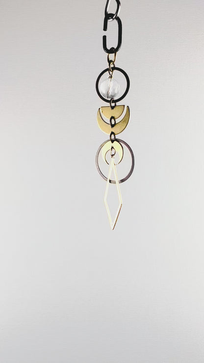 A slowly rotating piece of crystal rearview mirror art made up of connected gold colored metal components such as Rings, crescent moons, half moons and diamond shapes with a faceted clear quartz crystal bead hang in front of a white wall.