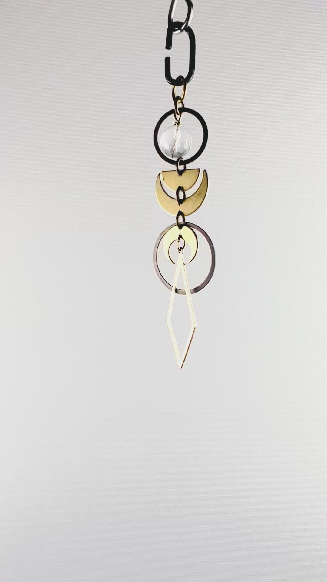 A slowly rotating piece of crystal rearview mirror art made up of connected gold colored metal components such as Rings, crescent moons, half moons and diamond shapes with a faceted clear quartz crystal bead hang in front of a white wall.