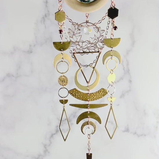 a piece of beautiful, crystal wall art with moss agate and garnet spins slowly in front of a gray and white marble wall. This hanging art includes connected parts such as: bronze colored chains, a light wood ring, a round moss agate stone. Connected to the wood, hanging gracefully below are gold and silver colored charms including: hexagons, half and crescent moons, rings, circles, diamonds and triangles; all connected by tiny rings.