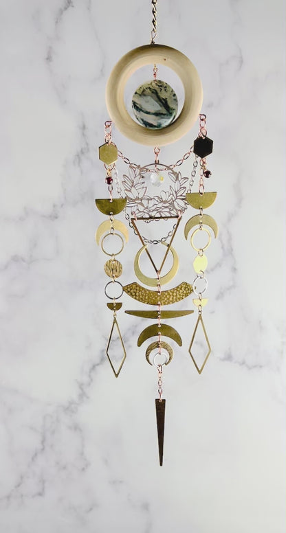a piece of beautiful, crystal wall art with moss agate and garnet spins slowly in front of a gray and white marble wall. This hanging art includes connected parts such as: bronze colored chains, a light wood ring, a round moss agate stone. Connected to the wood, hanging gracefully below are gold and silver colored charms including: hexagons, half and crescent moons, rings, circles, diamonds and triangles; all connected by tiny rings.