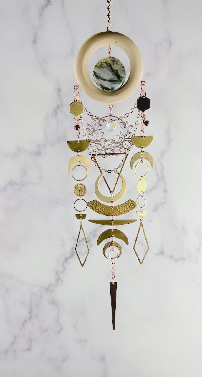 a piece of beautiful, crystal wall art with moss agate and garnet spins slowly in front of a gray and white marble wall. This hanging art includes connected parts such as: bronze colored chains, a light wood ring, a round moss agate stone. Connected to the wood, hanging gracefully below are gold and silver colored charms including: hexagons, half and crescent moons, rings, circles, diamonds and triangles; all connected by tiny rings.
