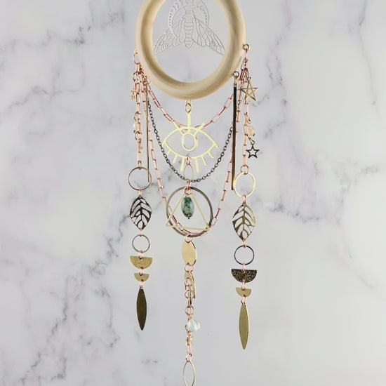 Hanging crystal wall art with Opalite Crescent Moon at the top, followed by a hammered arch and wood ring with Bee and moon phase in the middle. Hanging from the wood are copper and brass colored chains with an eye charm, stars, leaves, rings, half circles, circles, and long spears. The piece sparkles and rotates slowly in front of a marble wall.