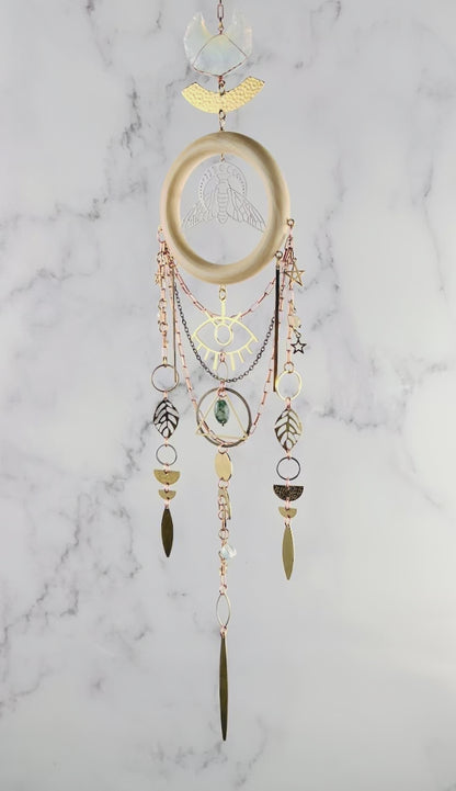 Hanging crystal wall art with Opalite Crescent Moon at the top, followed by a hammered arch and wood ring with Bee and moon phase in the middle. Hanging from the wood are copper and brass colored chains with an eye charm, stars, leaves, rings, half circles, circles, and long spears. The piece sparkles and rotates slowly in front of a marble wall.