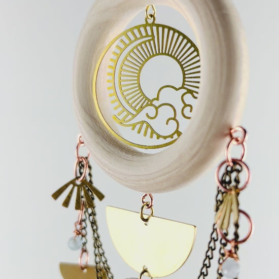 Aquamarine, green Tourmaline and Herkimer diamond crystal wall art slowly spins in front of a white wall. Elements include bronze chain, a light colored wood ring with gold metal sun, Moon, and cloud Charm in the center and dangling below are more bronze chains and three separate connected strand of gold colored metal shapes, such as half circles, moons, fans, crescent moons, a diamond, ovals, and a triangle at the bottom.
