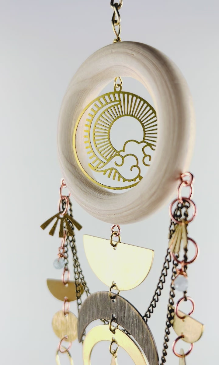 Aquamarine, green Tourmaline and Herkimer diamond crystal wall art slowly spins in front of a white wall. Elements include bronze chain, a light colored wood ring with gold metal sun, Moon, and cloud Charm in the center and dangling below are more bronze chains and three separate connected strand of gold colored metal shapes, such as half circles, moons, fans, crescent moons, a diamond, ovals, and a triangle at the bottom.