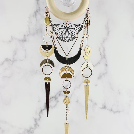 A hanging piece of crystal wall art JenA hanging piece of crystal wall, art gently spins as the camera zooms in and out. The peace includes a light wood ring with a round prism in the middle, hanging from a chain. Hanging from the ring are various metal pieces, including crescent, moons, rings, half circle, circles, triangle, wire, chains, wrapped beads, and a butterfly charm.