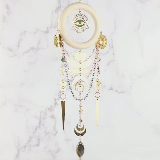 Hanging crystal wall art slowly spins in front of a marble wall. Featured on this are are a brass ring with natural Labradorite in the middle and below that a light wood ring hangs with an eye symbol surrounded by a triangle. Moss Agate stones arranged in a dimensional design, paired with gold colored metal, geometric shapes, and stars. The handcrafted piece measures approximately 16.5 inches tall and 4 inches wide.