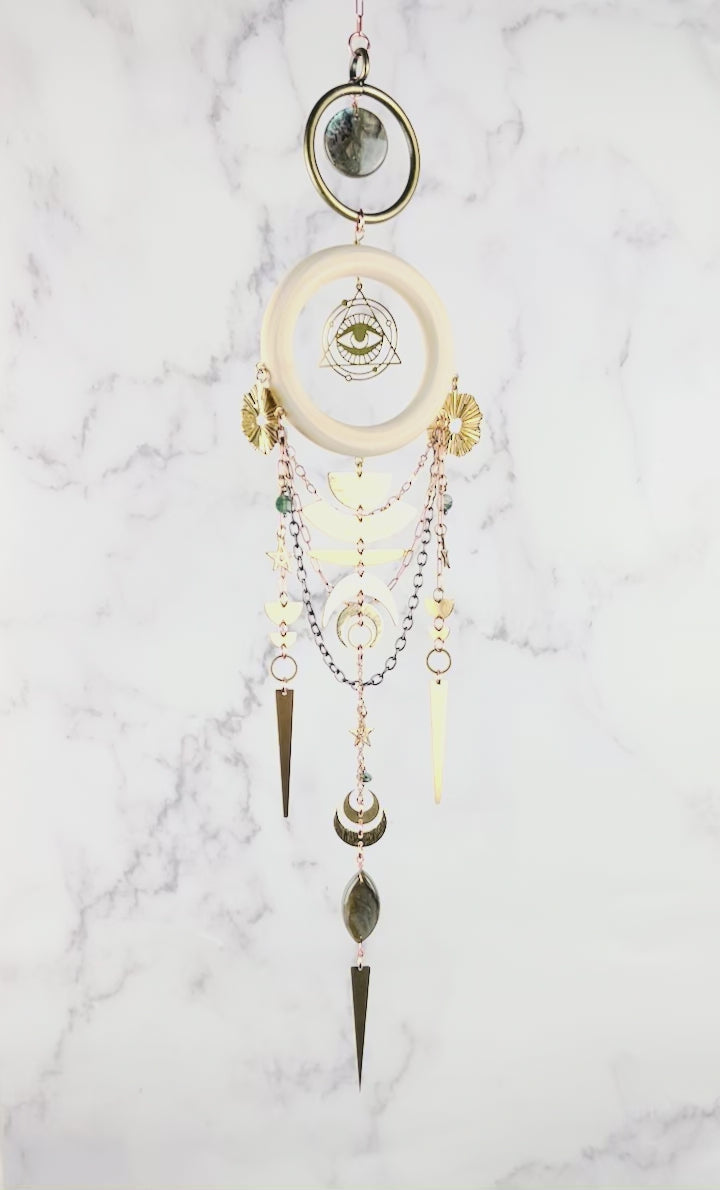 Hanging crystal wall art slowly spins in front of a marble wall. Featured on this are are a brass ring with natural Labradorite in the middle and below that a light wood ring hangs with an eye symbol surrounded by a triangle. Moss Agate stones arranged in a dimensional design, paired with gold colored metal, geometric shapes, and stars. The handcrafted piece measures approximately 16.5 inches tall and 4 inches wide.