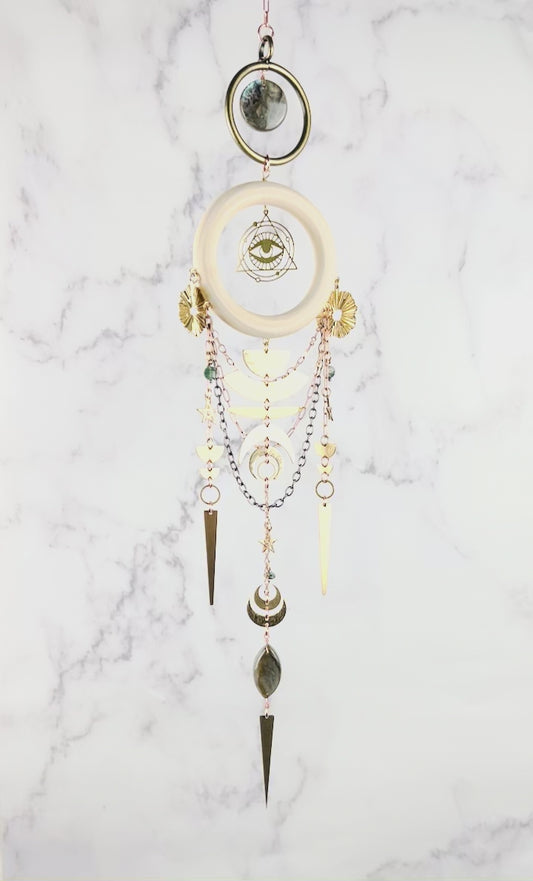 Hanging crystal wall art slowly spins in front of a marble wall. Featured on this are are a brass ring with natural Labradorite in the middle and below that a light wood ring hangs with an eye symbol surrounded by a triangle. Moss Agate stones arranged in a dimensional design, paired with gold colored metal, geometric shapes, and stars. The handcrafted piece measures approximately 16.5 inches tall and 4 inches wide.