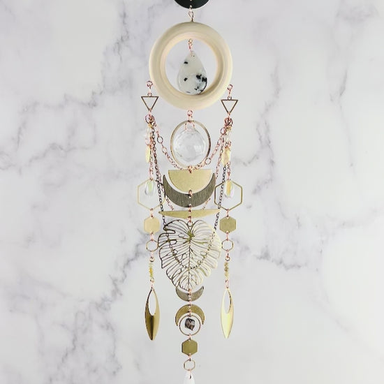 A slowly spinning, vertically hanging collage of wood, metal and crystals including: moonstone, selenite, green garnet, smoky quartz, and Seraphinite, create a beautiful piece of wall art. Included on this Charm are: copper colored chain, metal shaped such as hexagons, crescent, moons, triangles, half circles, ovals, Monstera leaves and ring, all hanging in front of a gray and white marble wall.