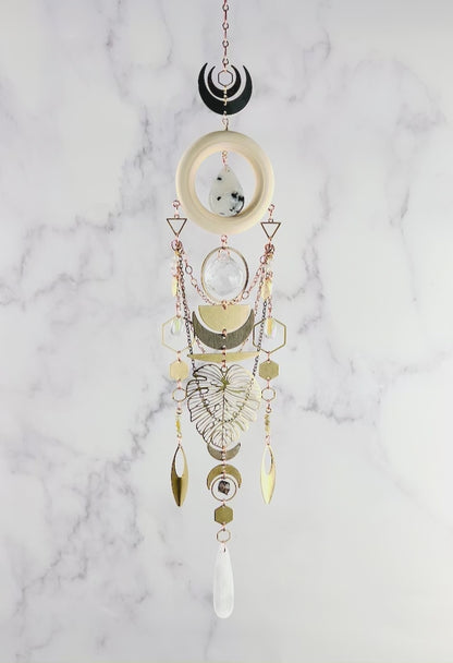 A slowly spinning, vertically hanging collage of wood, metal and crystals including: moonstone, selenite, green garnet, smoky quartz, and Seraphinite, create a beautiful piece of wall art. Included on this Charm are: copper colored chain, metal shaped such as hexagons, crescent, moons, triangles, half circles, ovals, Monstera leaves and ring, all hanging in front of a gray and white marble wall.