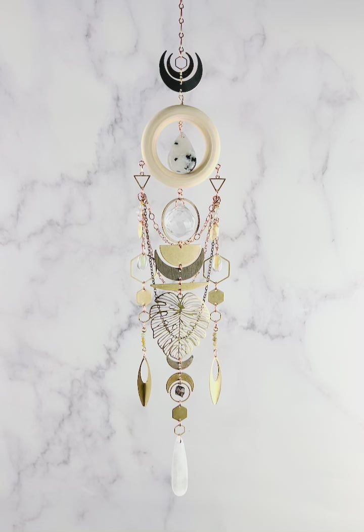 A slowly spinning, vertically hanging collage of wood, metal and crystals including: moonstone, selenite, green garnet, smoky quartz, and Seraphinite, create a beautiful piece of wall art. Included on this Charm are: copper colored chain, metal shaped such as hexagons, crescent, moons, triangles, half circles, ovals, Monstera leaves and ring, all hanging in front of a gray and white marble wall.