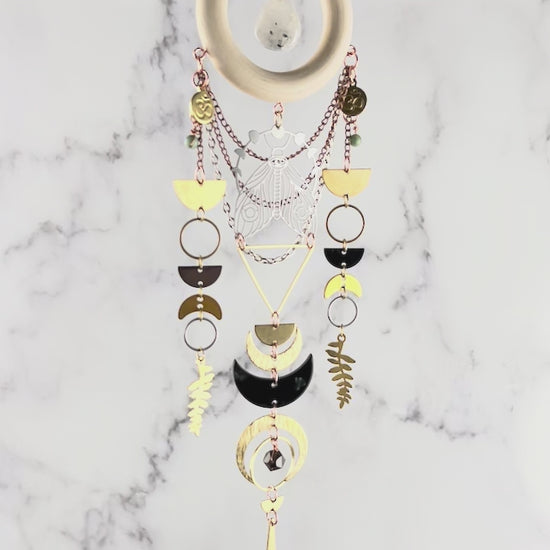 movie of a piece of moonstone, turquoise, quartz crystal wall art slowly rotating in front of a gray and white marble wall. Elements on the art include: a wood ring with a moonstone drop in the center, gold colored metal geometric shapes, Fern leaves, crescent moons, half circles and silver, and copper colored chain.