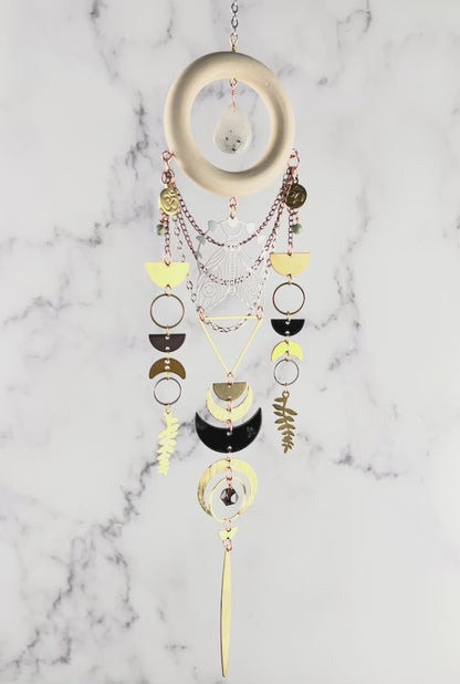 movie of a piece of moonstone, turquoise, quartz crystal wall art slowly rotating in front of a gray and white marble wall. Elements on the art include: a wood ring with a moonstone drop in the center, gold colored metal geometric shapes, Fern leaves, crescent moons, half circles and silver, and copper colored chain.