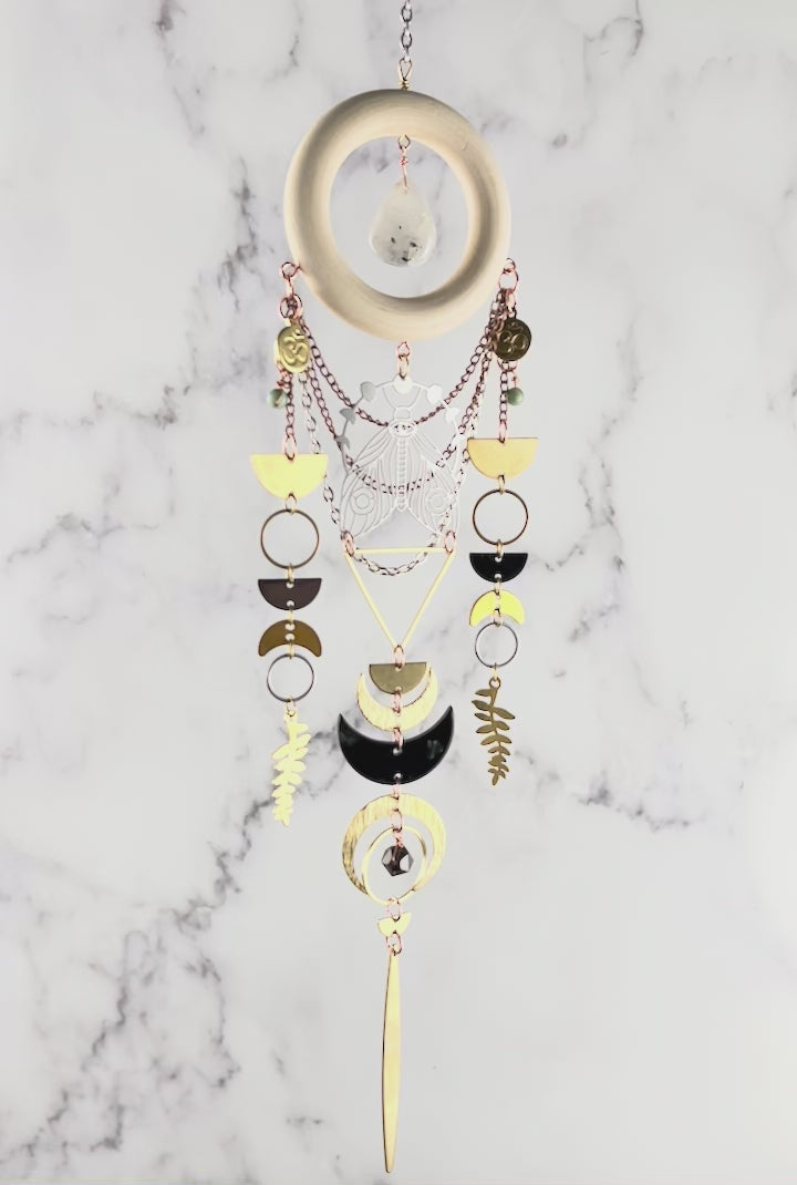 movie of a piece of moonstone, turquoise, quartz crystal wall art slowly rotating in front of a gray and white marble wall. Elements on the art include: a wood ring with a moonstone drop in the center, gold colored metal geometric shapes, Fern leaves, crescent moons, half circles and silver, and copper colored chain.