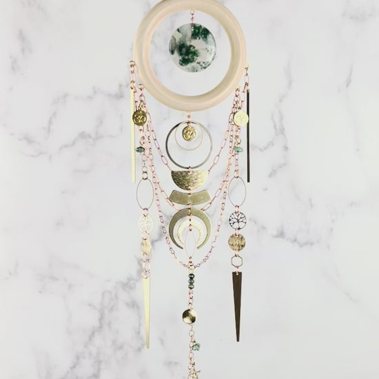 A piece of crystal wall art rotates slowly in front of a marble wall. The Charm features copper colored chain, crescent moons, a light wood ring with a piece of green and translucent disk of moss agate, at its center. Below hang more attached copper colored chains with connected gold colored metal tree of life, ohm symbol, wire wrapped beads, triangles, hexagons, ovals, stars, and a metal snake charm at the bottom.