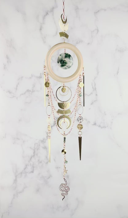 A piece of crystal wall art rotates slowly in front of a marble wall. The Charm features copper colored chain, crescent moons, a light wood ring with a piece of green and translucent disk of moss agate, at its center. Below hang more attached copper colored chains with connected gold colored metal tree of life, ohm symbol, wire wrapped beads, triangles, hexagons, ovals, stars, and a metal snake charm at the bottom.