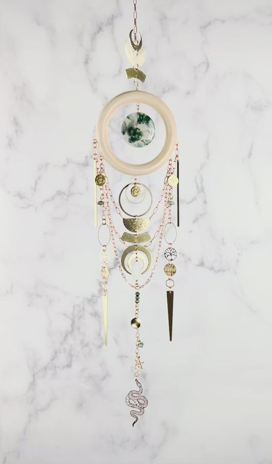 A piece of crystal wall art rotates slowly in front of a marble wall. The Charm features copper colored chain, crescent moons, a light wood ring with a piece of green and translucent disk of moss agate, at its center. Below hang more attached copper colored chains with connected gold colored metal tree of life, ohm symbol, wire wrapped beads, triangles, hexagons, ovals, stars, and a metal snake charm at the bottom.