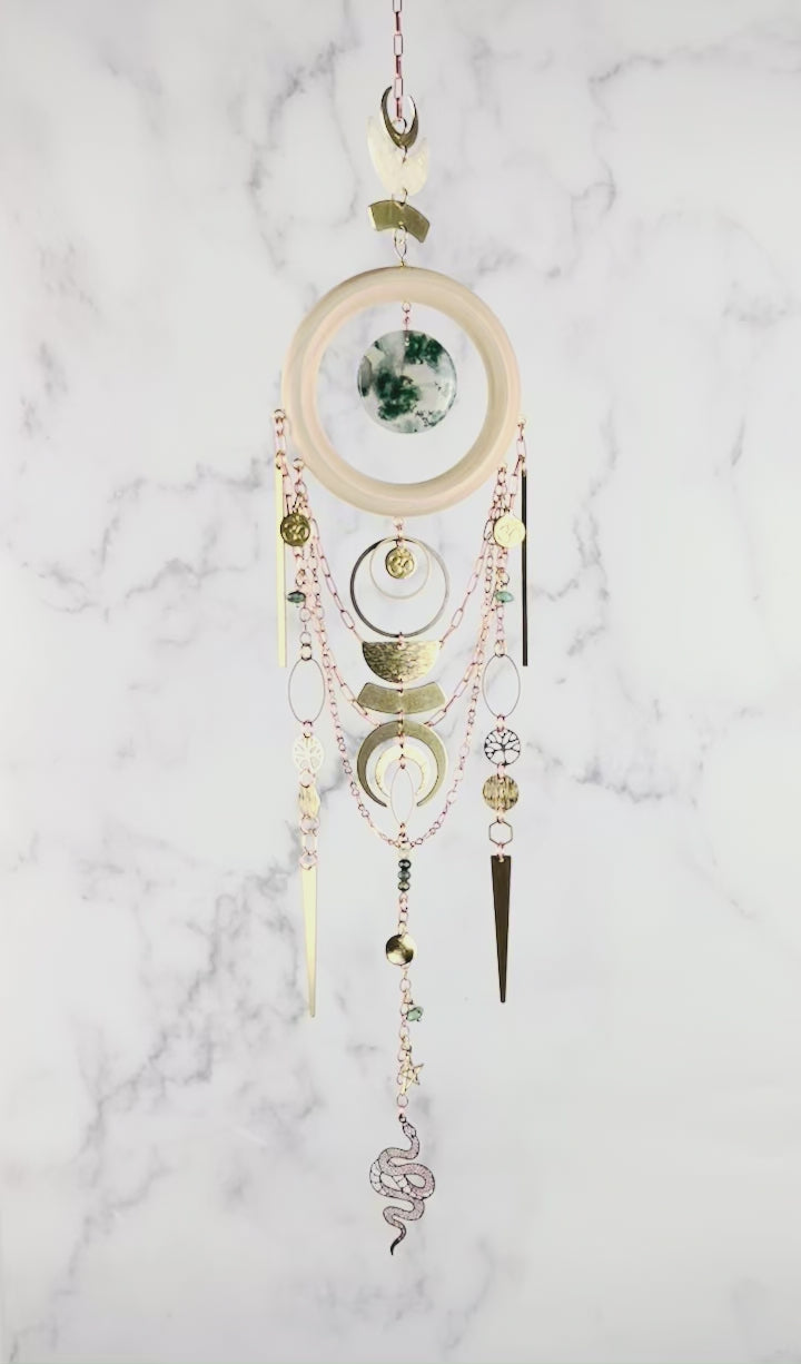 A piece of crystal wall art rotates slowly in front of a marble wall. The Charm features copper colored chain, crescent moons, a light wood ring with a piece of green and translucent disk of moss agate, at its center. Below hang more attached copper colored chains with connected gold colored metal tree of life, ohm symbol, wire wrapped beads, triangles, hexagons, ovals, stars, and a metal snake charm at the bottom.
