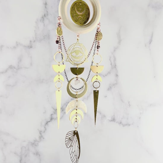 Celestial wall art with crystals, light, wood, ring, eye charm with triangle and Rings around it, bronze, colored chain, wire, wrapped beads, rings, half circles, crescent, moons, citrine, prisms, and a bug wing hanging from a chain and slowly rotating in front of a marble wall.