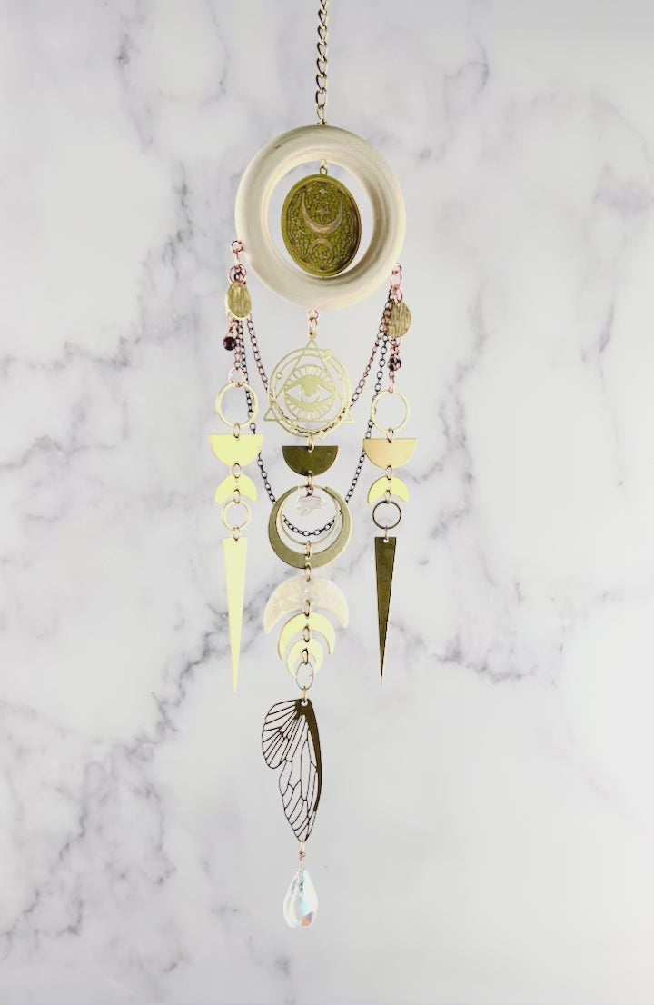 Celestial wall art with crystals, light, wood, ring, eye charm with triangle and Rings around it, bronze, colored chain, wire, wrapped beads, rings, half circles, crescent, moons, citrine, prisms, and a bug wing hanging from a chain and slowly rotating in front of a marble wall.