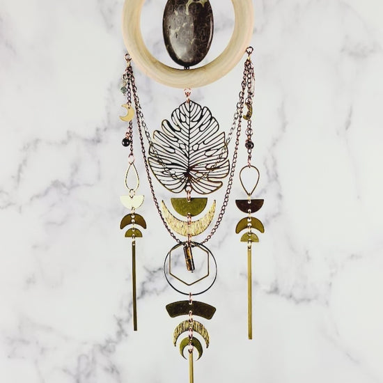 Hanging crystal wall art with gold and silver colored chain, Crescent moons, a large Monstera metal Charm, half circles, teardrop shapes, bars, hexagons, and rings all in gold and silver metals, slowly spins in front of a marble background.