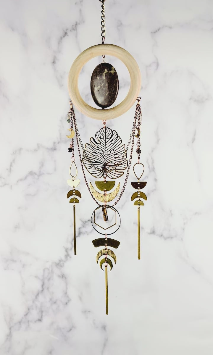 Hanging crystal wall art with gold and silver colored chain, Crescent moons, a large Monstera metal Charm, half circles, teardrop shapes, bars, hexagons, and rings all in gold and silver metals, slowly spins in front of a marble background.