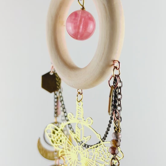 A piece of crystal wall art with cherry quartz, rhodochrosite, and pink aventurine spins gently in front of a light gray wall. Components on this vertically hanging art include: gold colored metal hexagons, a moth charm, with sword, triangles, bronze, bells, silver rings, wire, wrapped beads, bronze, and silver chain, half moons, crescent, moons, and long gold bars.