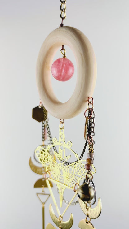 A piece of crystal wall art with cherry quartz, rhodochrosite, and pink aventurine spins gently in front of a light gray wall. Components on this vertically hanging art include: gold colored metal hexagons, a moth charm, with sword, triangles, bronze, bells, silver rings, wire, wrapped beads, bronze, and silver chain, half moons, crescent, moons, and long gold bars.