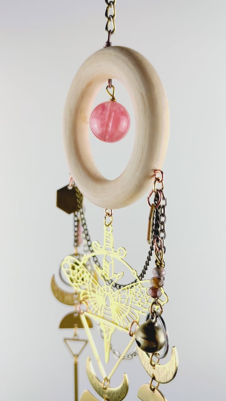 A piece of crystal wall art with cherry quartz, rhodochrosite, and pink aventurine spins gently in front of a light gray wall. Components on this vertically hanging art include: gold colored metal hexagons, a moth charm, with sword, triangles, bronze, bells, silver rings, wire, wrapped beads, bronze, and silver chain, half moons, crescent, moons, and long gold bars.