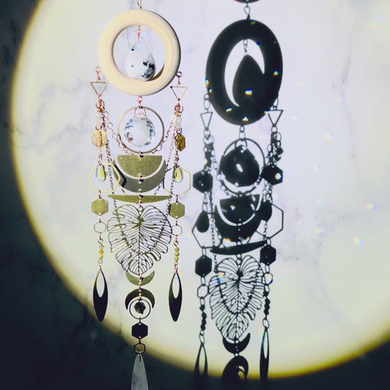 A crystal wall art with moonstone, prism, selenite, green garnet, and smoky quartz spins gently and sparkles in a spotlight. A cream colored wood ring with moonstone drop crystal hangs in its center. Attached to the wood, hanging vertically below it are beautiful elements include: copper colored chain, various silver, and gold charms, including crescent moons, hexagons, triangles, rings, ovals, a large Monstera leaf, all connected with tiny rings.