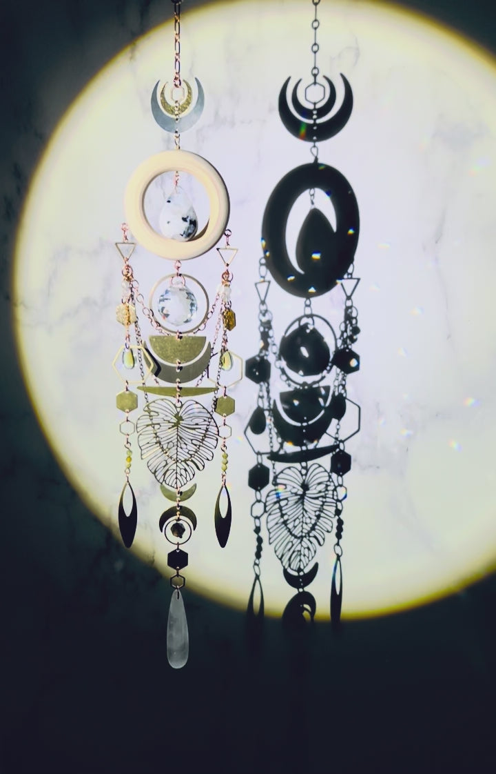 A crystal wall art with moonstone, prism, selenite, green garnet, and smoky quartz spins gently and sparkles in a spotlight. A cream colored wood ring with moonstone drop crystal hangs in its center. Attached to the wood, hanging vertically below it are beautiful elements include: copper colored chain, various silver, and gold charms, including crescent moons, hexagons, triangles, rings, ovals, a large Monstera leaf, all connected with tiny rings.