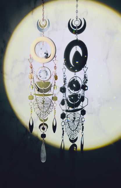 A crystal wall art with moonstone, prism, selenite, green garnet, and smoky quartz spins gently and sparkles in a spotlight. A cream colored wood ring with moonstone drop crystal hangs in its center. Attached to the wood, hanging vertically below it are beautiful elements include: copper colored chain, various silver, and gold charms, including crescent moons, hexagons, triangles, rings, ovals, a large Monstera leaf, all connected with tiny rings.