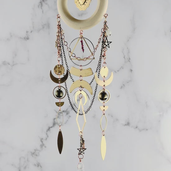 Red Garnet and pink adventurine crystal wall art with light wood ring and heart Charm in the center. Chains hang from the wood ring, with other shapes, such as stars, crescent moons, circles, rings, diamonds, and other various wire wrapped beads with a prism at the bottom. This piece is turning gently in front of a marble wall.