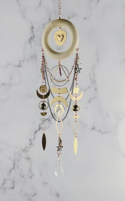 Red Garnet and pink adventurine crystal wall art with light wood ring and heart Charm in the center. Chains hang from the wood ring, with other shapes, such as stars, crescent moons, circles, rings, diamonds, and other various wire wrapped beads with a prism at the bottom. This piece is turning gently in front of a marble wall.