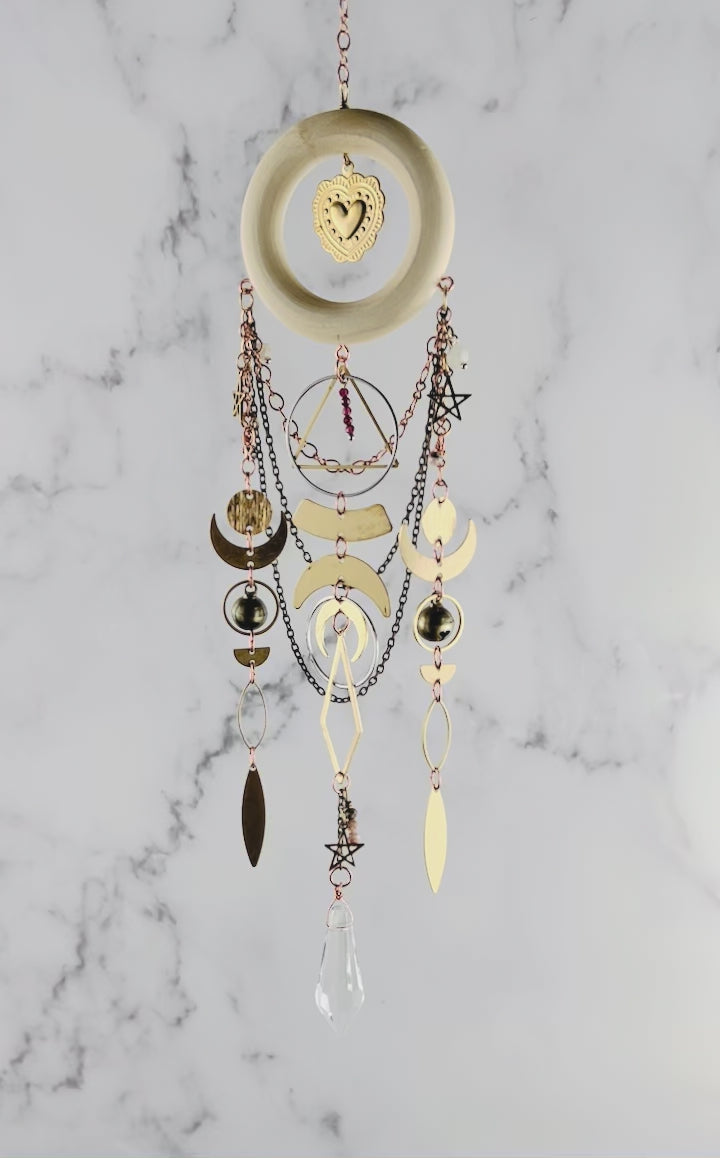 Red Garnet and pink adventurine crystal wall art with light wood ring and heart Charm in the center. Chains hang from the wood ring, with other shapes, such as stars, crescent moons, circles, rings, diamonds, and other various wire wrapped beads with a prism at the bottom. This piece is turning gently in front of a marble wall.