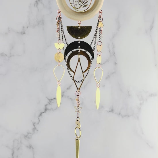 Piece of vertical crystal wall art gently spins in front of a marble wall. The piece consists of gold colored chain, a wood ring with a sun and clouds Charm in the middle, various other metal shapes on the charm include: half circles, circles, upside down teardrop shapes, a diamond, star, citrine, moonstone, half circles, triangle, and crescent moons.
