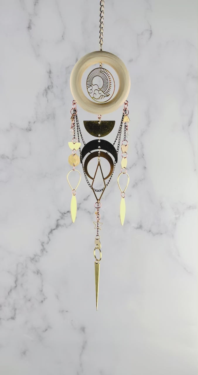 Piece of vertical crystal wall art gently spins in front of a marble wall. The piece consists of gold colored chain, a wood ring with a sun and clouds Charm in the middle, various other metal shapes on the charm include: half circles, circles, upside down teardrop shapes, a diamond, star, citrine, moonstone, half circles, triangle, and crescent moons.