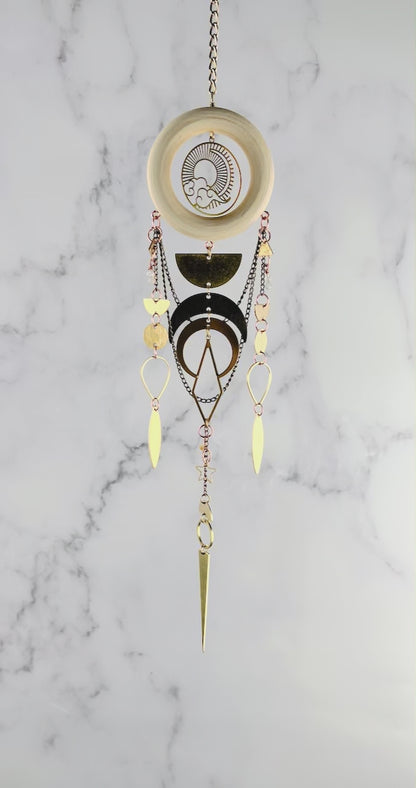 Piece of vertical crystal wall art gently spins in front of a marble wall. The piece consists of gold colored chain, a wood ring with a sun and clouds Charm in the middle, various other metal shapes on the charm include: half circles, circles, upside down teardrop shapes, a diamond, star, citrine, moonstone, half circles, triangle, and crescent moons.