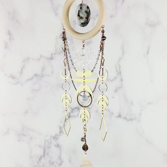 A piece of Bohemian style crystal wall art slowly spins in front of a gray and white marble background. The piece includes connected geometric and natural Charms a moon theme and chains with a snake Charm at the very bottom. The piece resembles a Dreamcatcher, made of mixed natural materials.