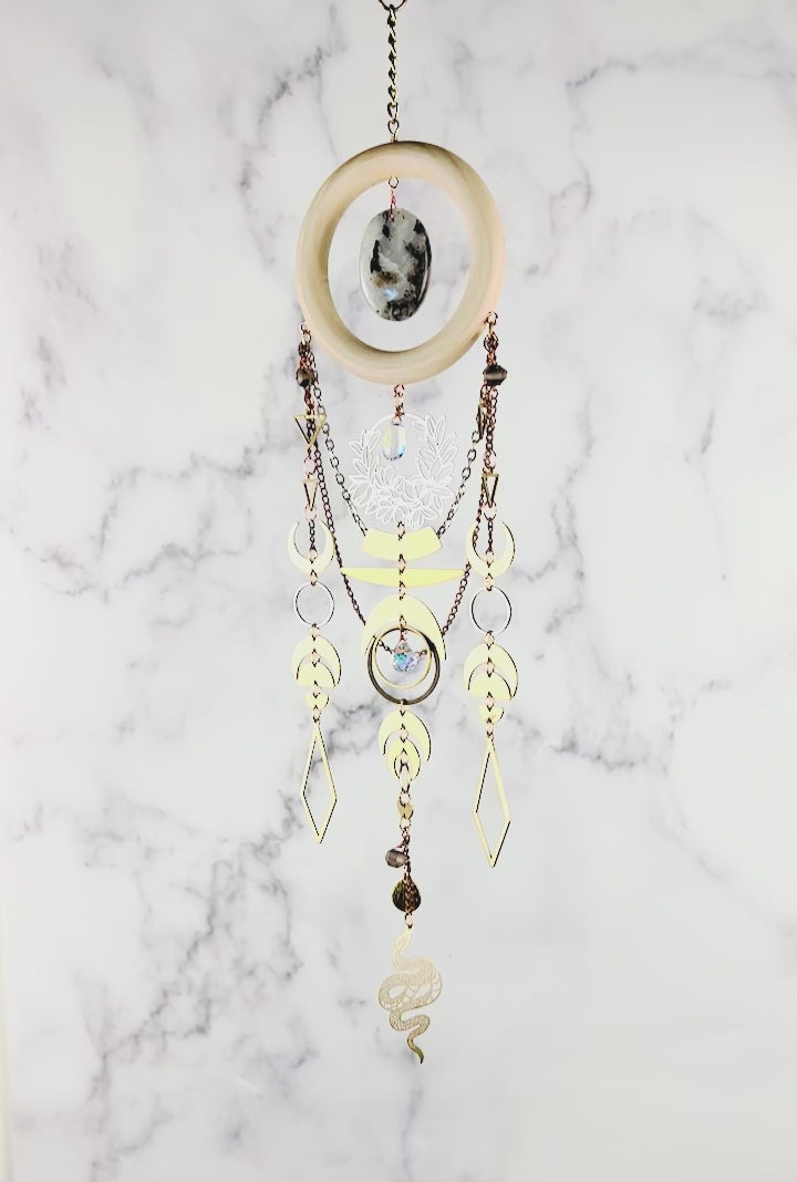 A piece of Bohemian style crystal wall art slowly spins in front of a gray and white marble background. The piece includes connected geometric and natural Charms a moon theme and chains with a snake Charm at the very bottom. The piece resembles a Dreamcatcher, made of mixed natural materials.