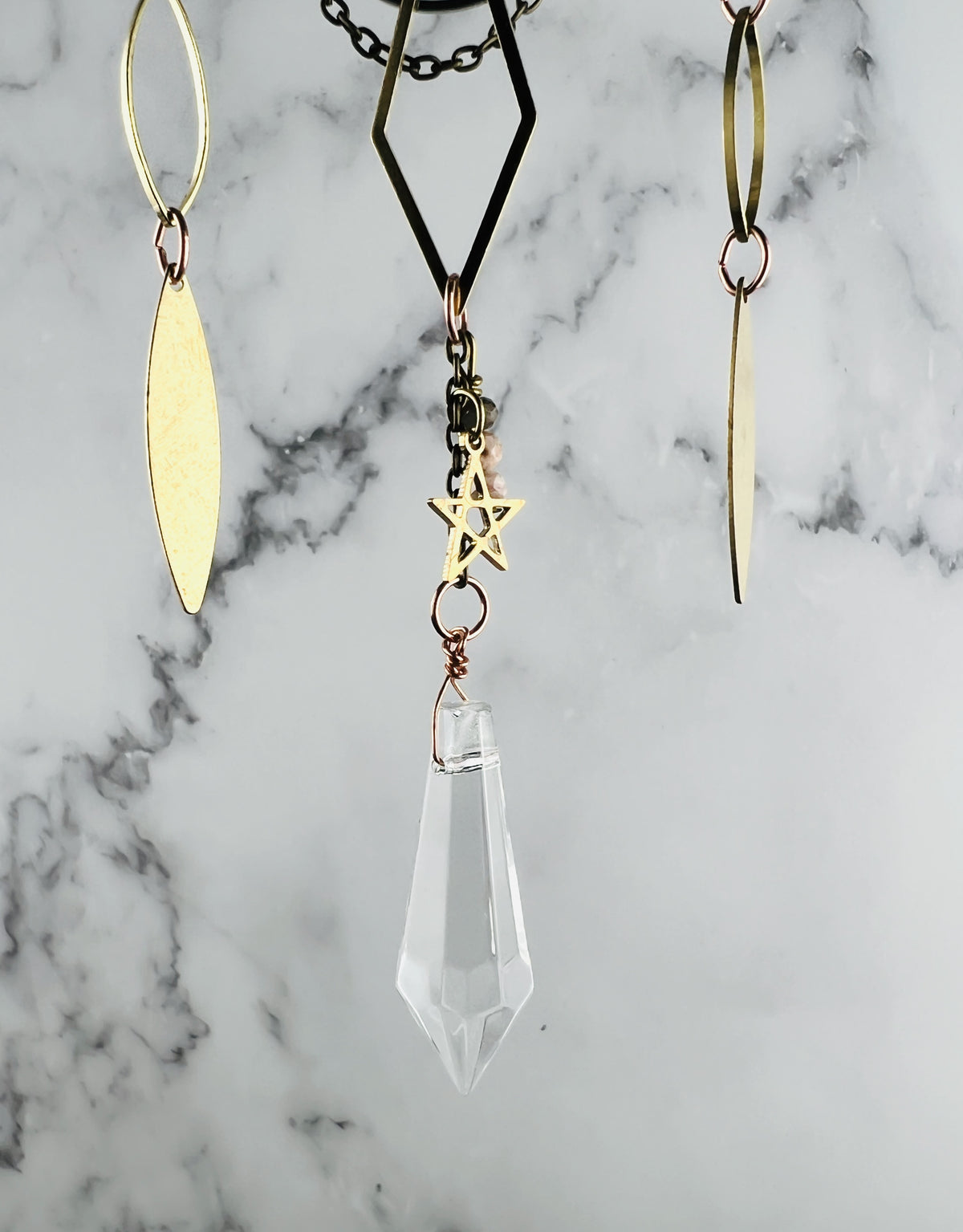 A gold-toned metal mobile with three hanging charms. The charms are a diamond shape, a drop shape, and a star and wire wrapped crystal prism hanging from the bottom. It hangs in front of a marble wall.