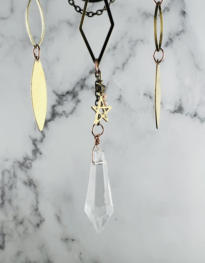 A gold-toned metal mobile with three hanging charms. The charms are a diamond shape, a drop shape, and a star and wire wrapped crystal prism hanging from the bottom. It hangs in front of a marble wall.