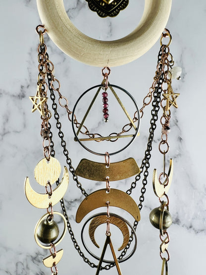 Hanging wooden ring at the top with gold charm in the middle. The crystal wall art features multiple gold colored crescent moon shapes, a triangle, and stars. Attached are multiple chains, rings, bells and half circles. It hangs in front of a marble wall.