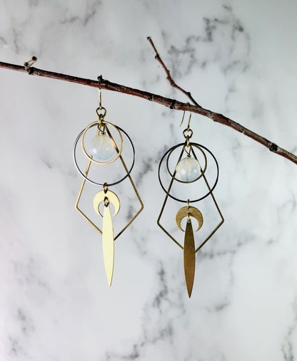 Picture shows pair of faceted Opalite dangle earrings with moon theme and reversed metal diamond component. Also include included on these earrings are steel and gold, colored rings, crescent moon, and long pointed ovals. They hang in front of a marble wall on a thin branch.
