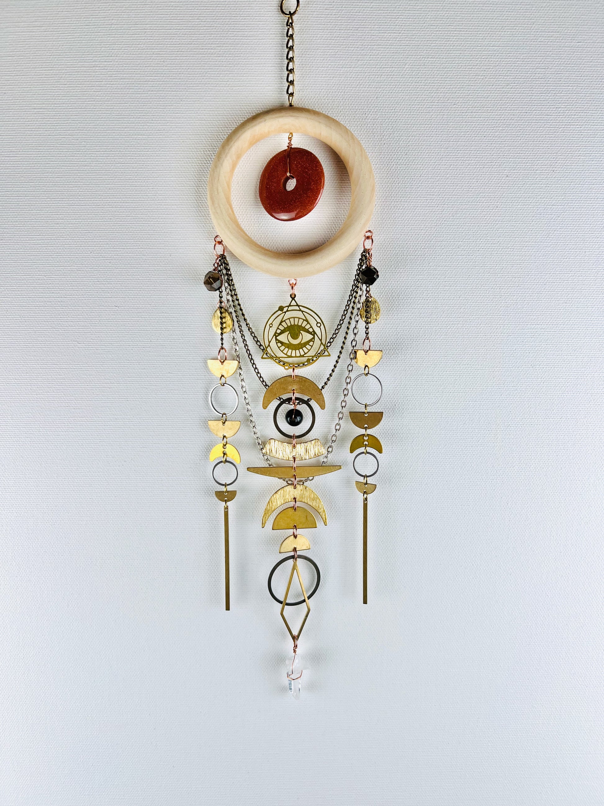 Decorative hanging crystal wall art with sandstone, smoky quartz, and clear quartz with various gold and silver charms, including, the evil eye protection symbol, half circles, rings, drops, sticks, diamonds, and crescent moons with a wood ring and chain hanging from it, on a light gray background