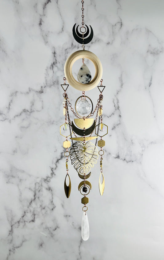 Featuring a beautifully designed piece of three-dimensional hanging wall art including a copper colored chain, followed vertically by a small hexagon, surrounded by crescent moons, below that hangs a cream colored wood ring with a moonstone hanging in the center. Attached to that are metal shapes and crystals, including: triangles, rings, copper colored chain, bronze colored chain, wire wrapped beads, half circles, hexagons, ovals, and a large Monstera leaf, hanging in front of a gray and white marble wall