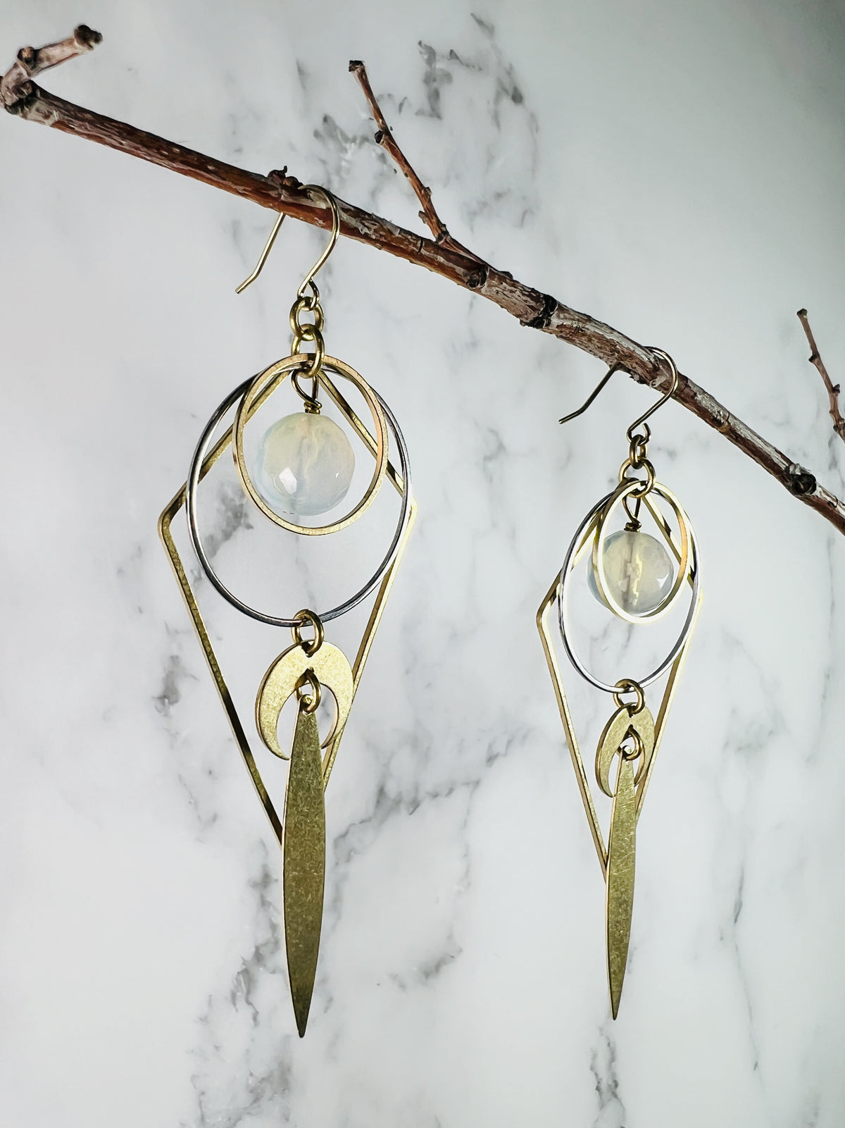 Close-up, angle side view of cited Opalite crystal earrings that dangle from a thin branch in front of a marble wall. Other components on these earrings include silver and gold, colored metal rings, Crescent moons, long pointed ovals and steel ear hooks.